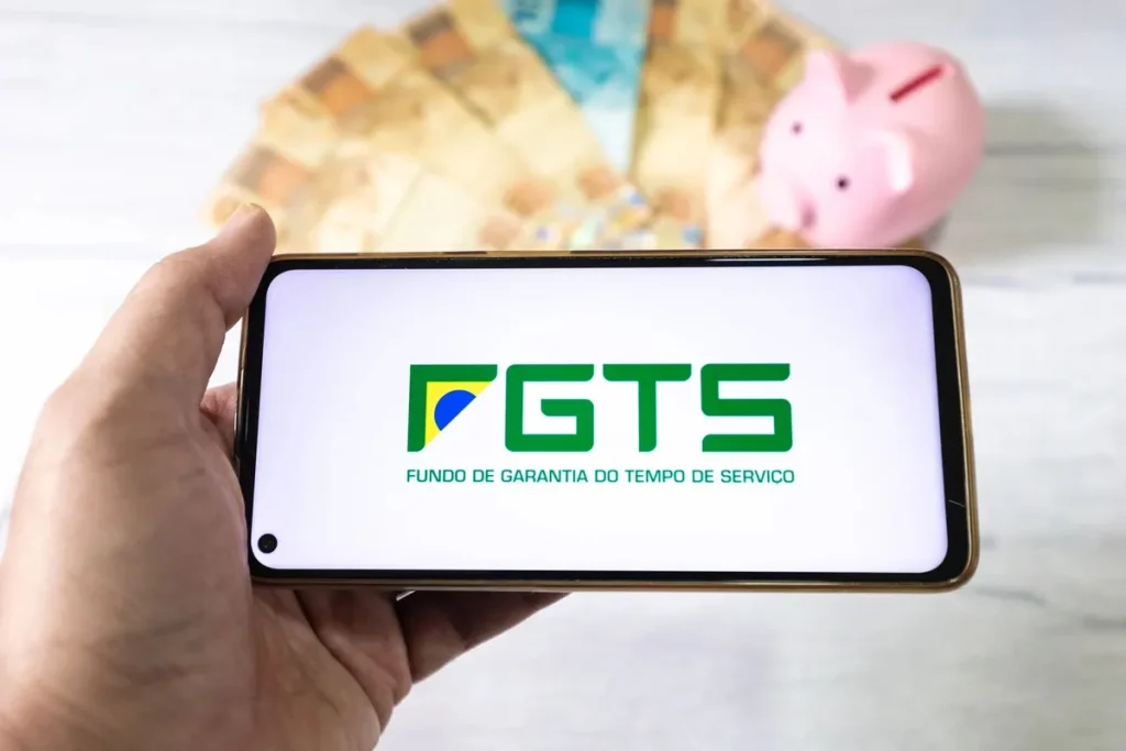 Tela de celular exibindo o logotipo do FGTS com notas de dinheiro e cofre em formato de porquinho ao fundo