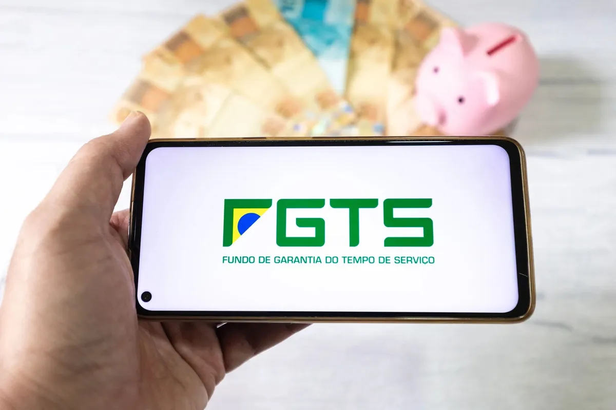 Tela de celular exibindo o logotipo do FGTS com notas de dinheiro e cofre em formato de porquinho ao fundo