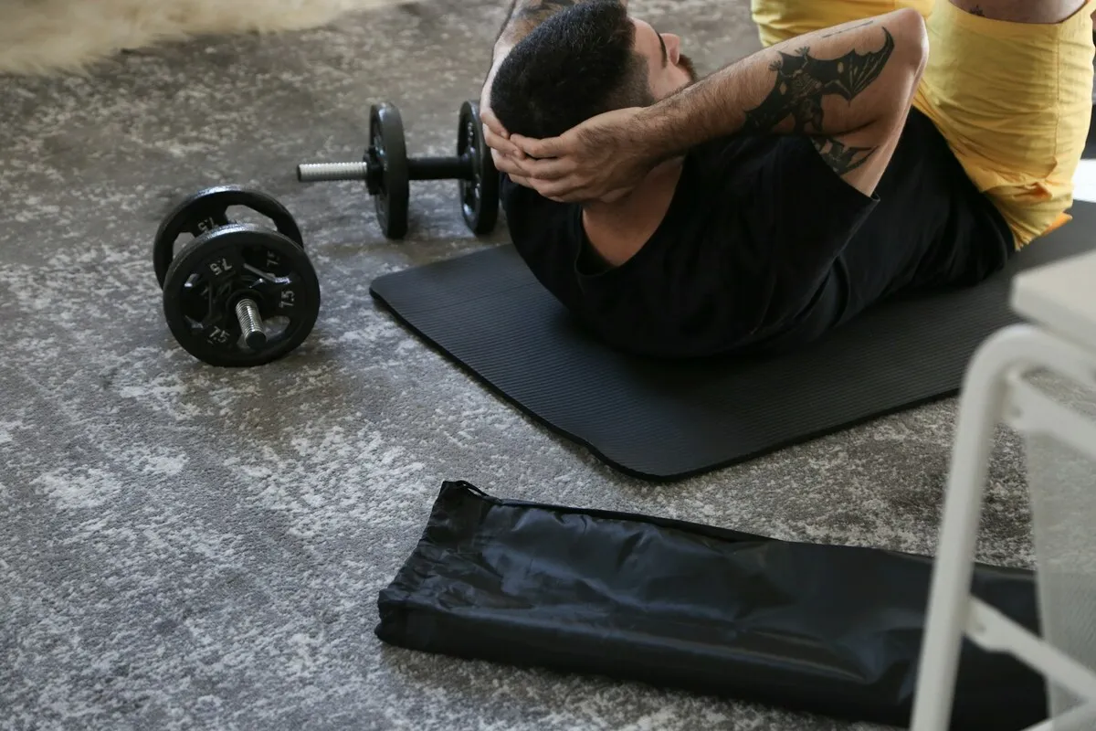 Homem fazendo abdominais em tapete de yoga preto, com halteres ao lado, durante treino em casa.