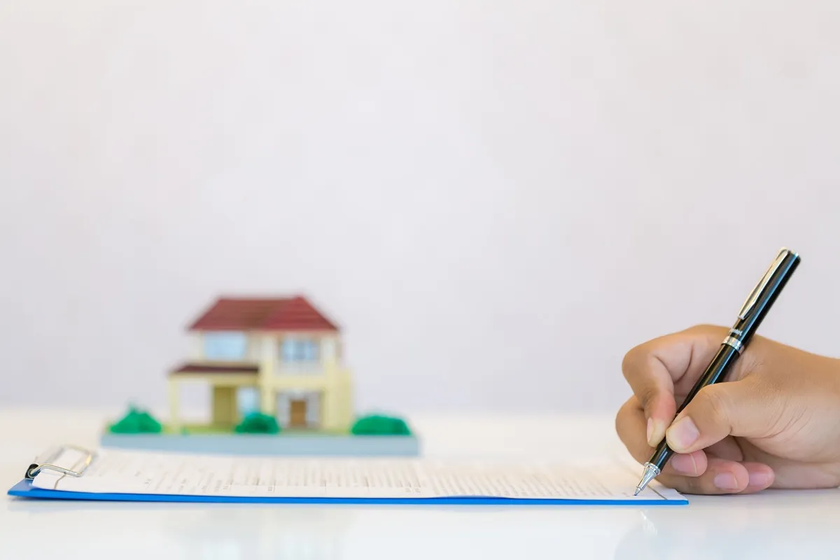 Imagem mostrando uma mão assinando a escritura de um imóvel com uma caneta, enquanto ao fundo aparece um modelo de casa de brinquedo