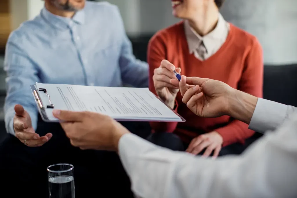 Uma imagem em close-up mostra um casal sorrindo e prestes a assinar um documento em uma prancheta. Um consultor financeiro ou advogado, cujas mãos seguram a prancheta e oferecem a caneta, guia o casal no processo. O ambiente é formal, e a linguagem corporal do casal, que parece aliviado e feliz, sugere que eles estão finalizando um acordo importante ou uma negociação de dívidas bem-sucedida. A cena representa a solução para a inadimplência, simbolizando o momento de sair das dívidas e conquistar a estabilidade financeira por meio de um acordo formal e estruturado.