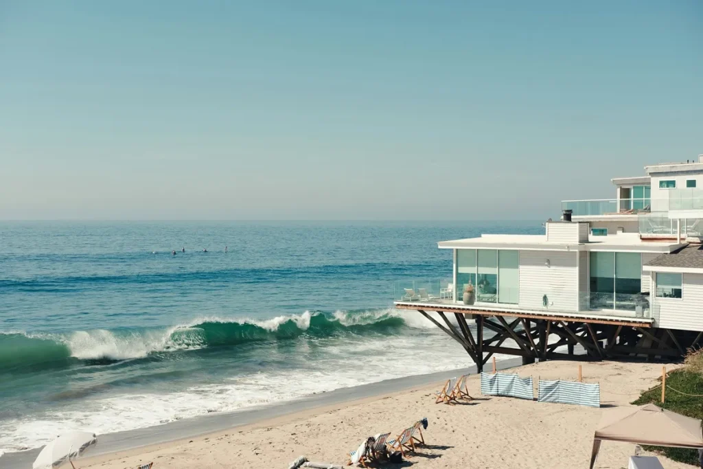 Casa de praia moderna elevada sobre estacas, com grande varanda de vidro e vista para o oceano, enquanto ondas quebram na areia e pessoas praticam surfe