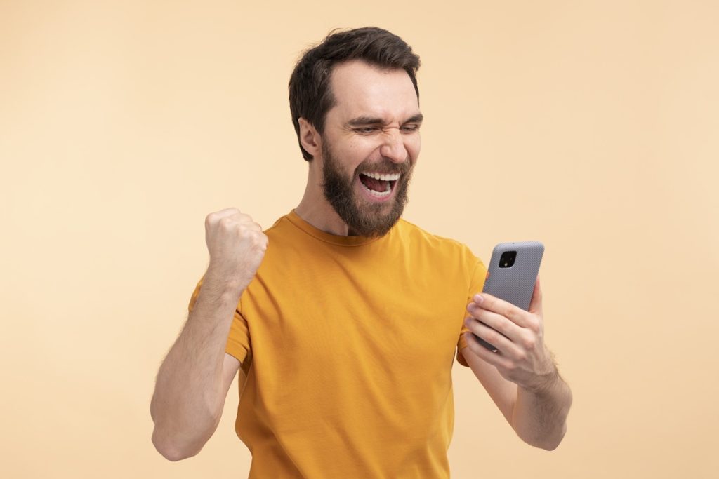 Homem sorrindo e levantando o punho em vitória enquanto olha para o celular, com fundo de cor suave.