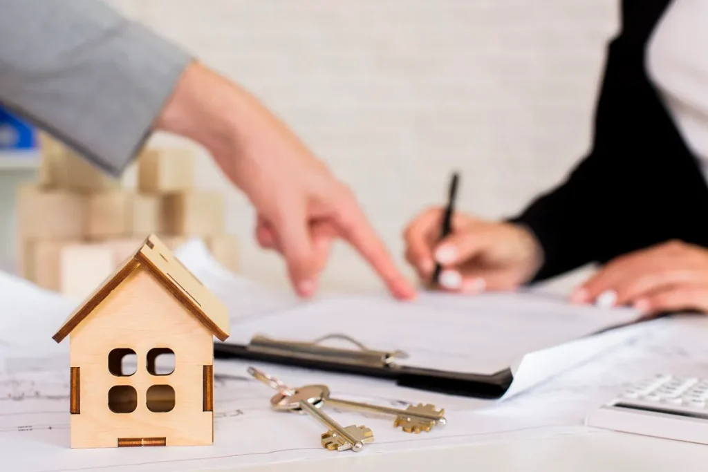 Imagem mostrando a assinatura de escritura de imóvel, com uma mão apontando para o contrato e outra pessoa assinando, enquanto ao fundo há uma miniatura de casa de madeira e chaves.