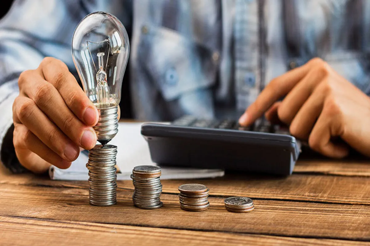 Pessoa segurando lâmpada sobre moedas empilhadas ao lado de calculadora, simbolizando economia e planejamento financeiro