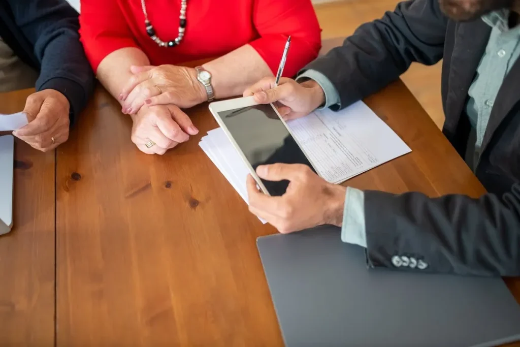 Três pessoas em uma mesa de madeira, com um homem de terno apontando para um tablet, representando uma reunião de negócios ou uma assembleia para tomada de decisões.