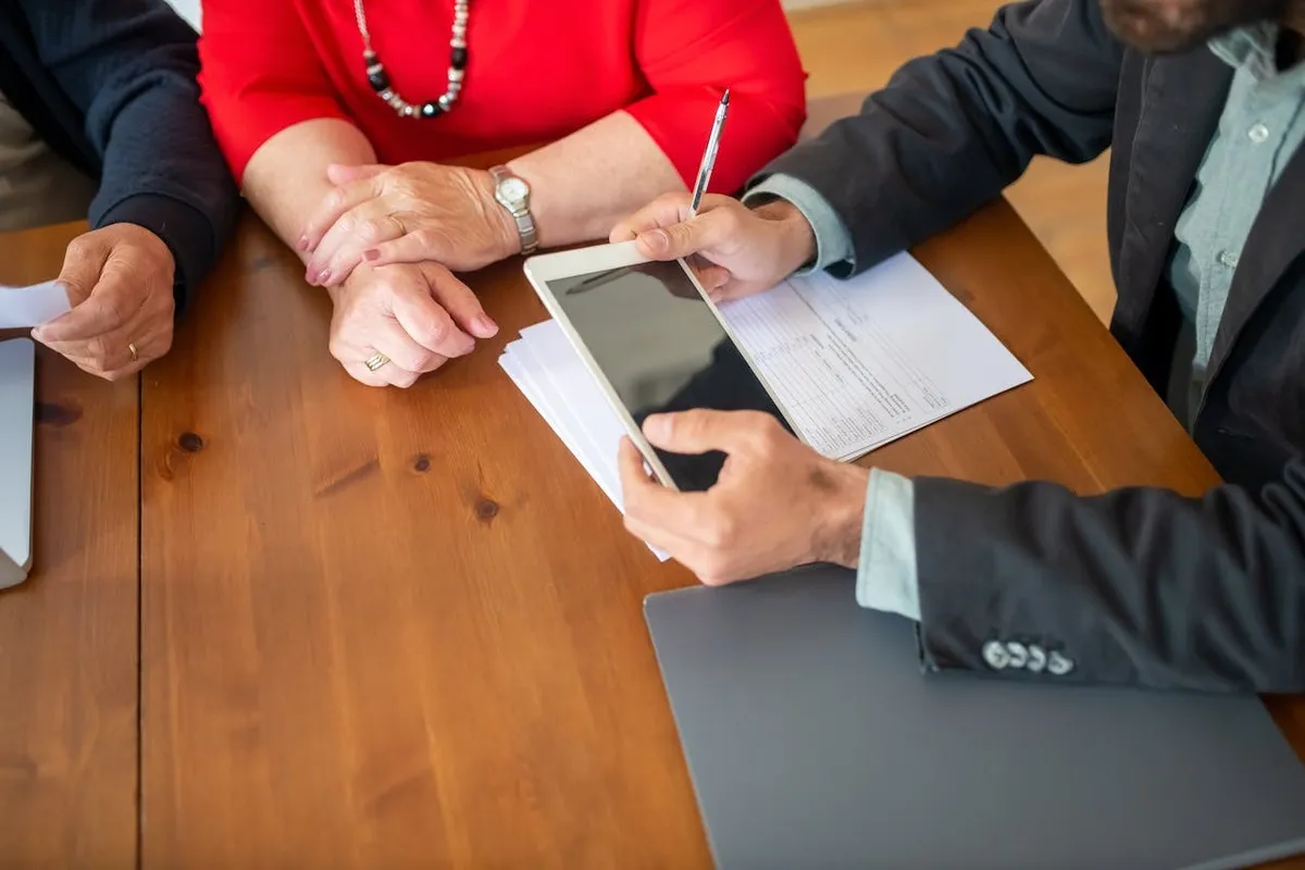 Três pessoas em uma mesa de madeira, com um homem de terno apontando para um tablet, representando uma reunião de negócios ou uma assembleia para tomada de decisões.