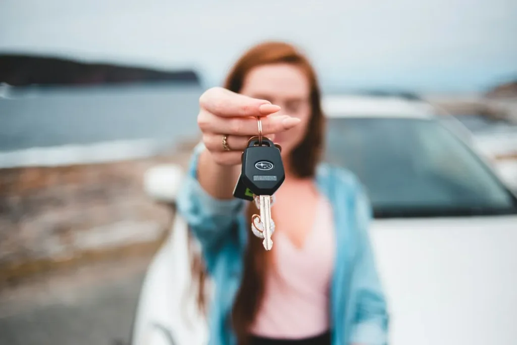 Uma pessoa com cabelo ruivo desfocado segura um chaveiro da Subaru com as chaves do carro em primeiro plano, com um carro branco e um lago ou mar ao fundo, simbolizando a aquisição de um veículo.