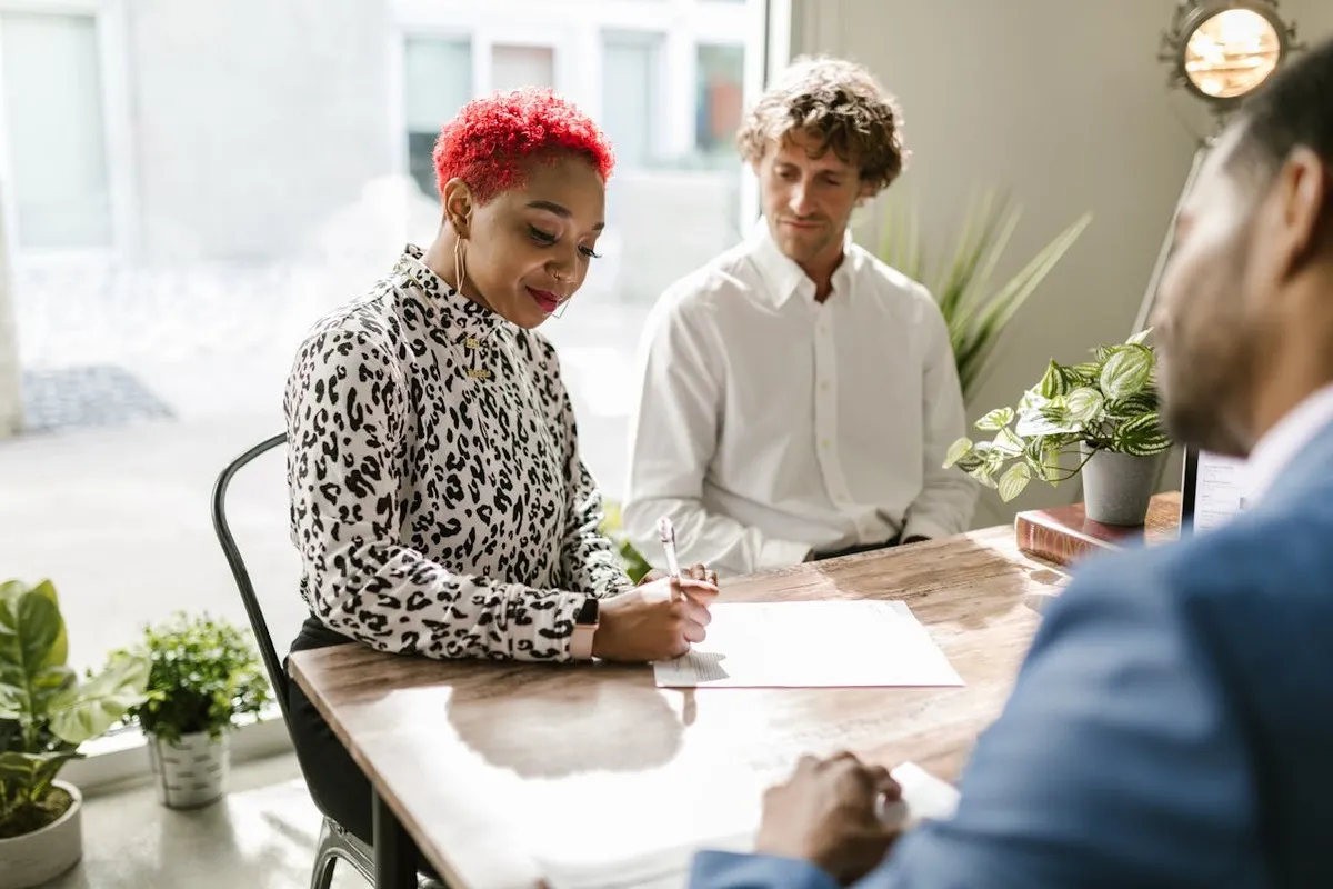 Uma mulher com cabelo vermelho vibrante assinando um documento em uma mesa de madeira, enquanto dois homens a observam, um deles de costas, em um ambiente de reunião ou escritório bem iluminado.