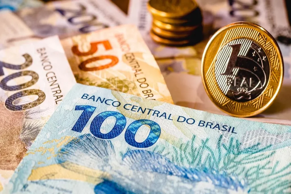 Cédulas de 100 e 50 reais com uma moeda de 1 real em primeiro plano, sobre um fundo de outras notas e moedas.