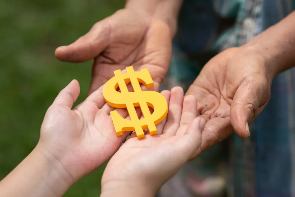 Mãos de uma pessoa adulta entregando um símbolo de dólar amarelo para as mãos de uma pessoa mais jovem, em um gesto de apoio financeiro, ilustrando a palavra-chave 'empréstimo pessoal'.