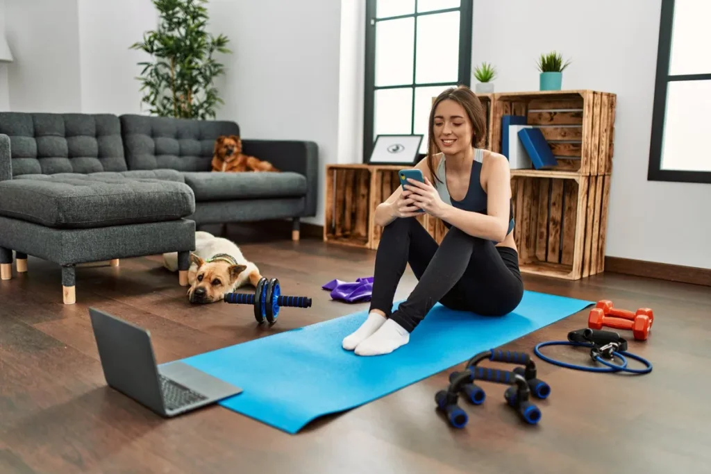 Uma mulher sentada em um tapete de ioga azul, olhando para o celular, com vários equipamentos de academia ao seu redor, como halteres, uma corda e um rolo abdominal. Seus dois cachorros estão deitados no chão e no sofá.