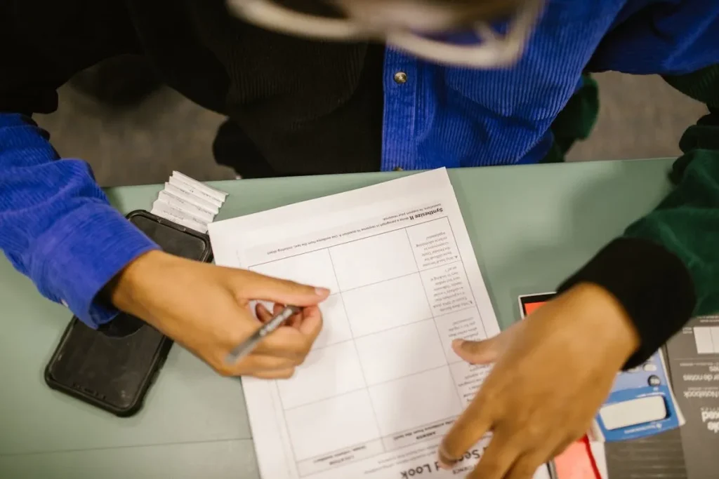 Pessoa preenchendo formulário com caneta e celular ao lado, representando o processo de planejamento para financiar faculdade.