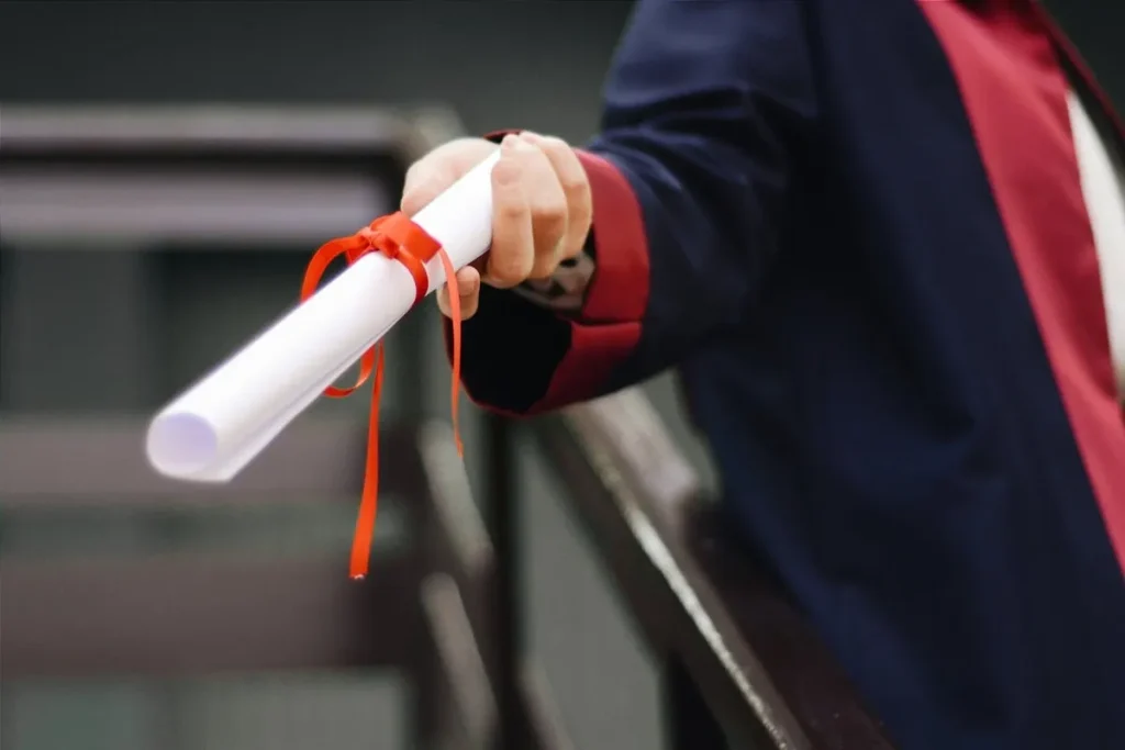 Mão segurando diploma enrolado e amarrado com fita vermelha, simbolizando a conclusão da faculdade e o investimento em educação.