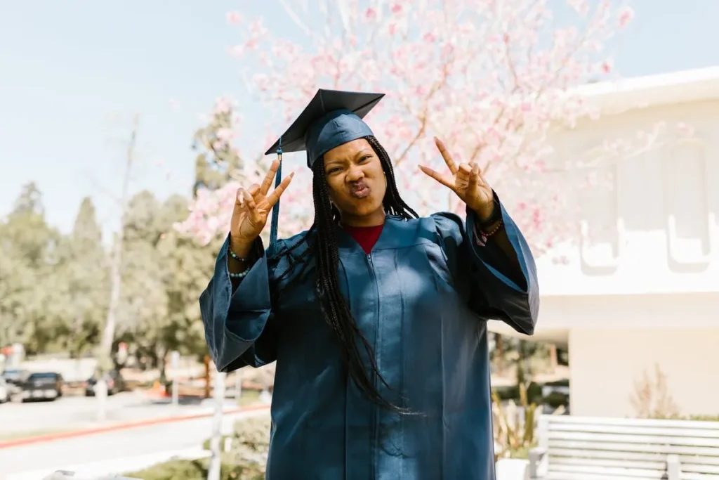 Jovem mulher negra sorridente vestindo beca e capelo de formatura, fazendo sinal de paz com as mãos, celebrando a conquista de financiar a faculdade.