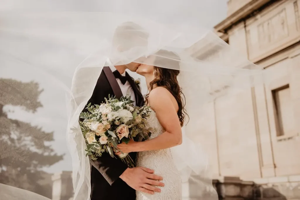 Um casal em trajes de casamento se beija, com seus rostos parcialmente cobertos por um véu translúcido. A noiva segura um buquê de flores brancas e rosadas. Ao fundo, um grande edifício de pedra e árvores.