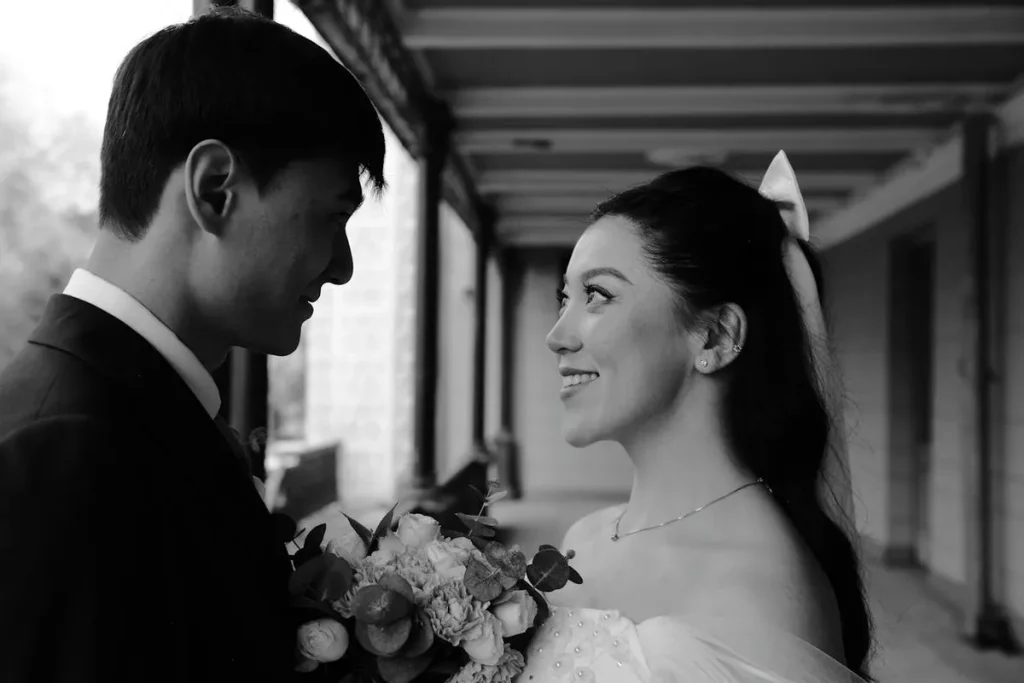 Uma fotografia em preto e branco de um casal em trajes de casamento, olhando um para o outro e sorrindo. O noivo, de terno, e a noiva, com um vestido tomara que caia, seguram um buquê e estão em um corredor.