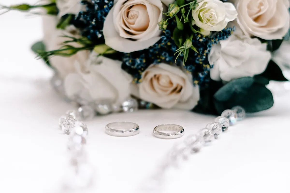 Close-up de duas alianças de casamento de prata ou platina, dispostas em frente a um buquê de noiva com rosas brancas e um terço de cristal, em uma superfície branca.
