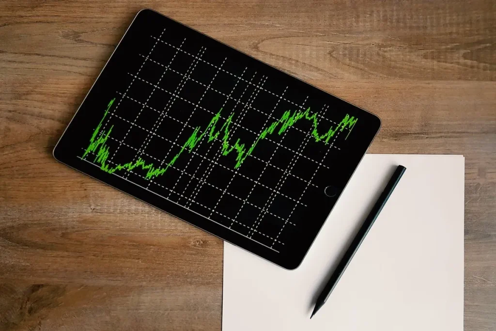 Uma mão segurando um tablet com um gráfico de linha verde, que mostra o aumento de valores ao longo do tempo. Um lápis preto e uma folha de papel branca estão ao lado, em uma mesa de madeira.