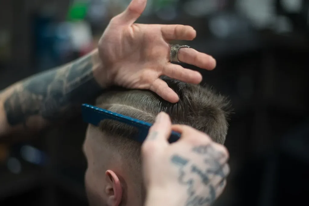 Um barbeiro com tatuagens nos braços, segurando um pente azul, corta o cabelo de um cliente. A imagem foca no trabalho manual e na especialização dos profissionais autônomos.