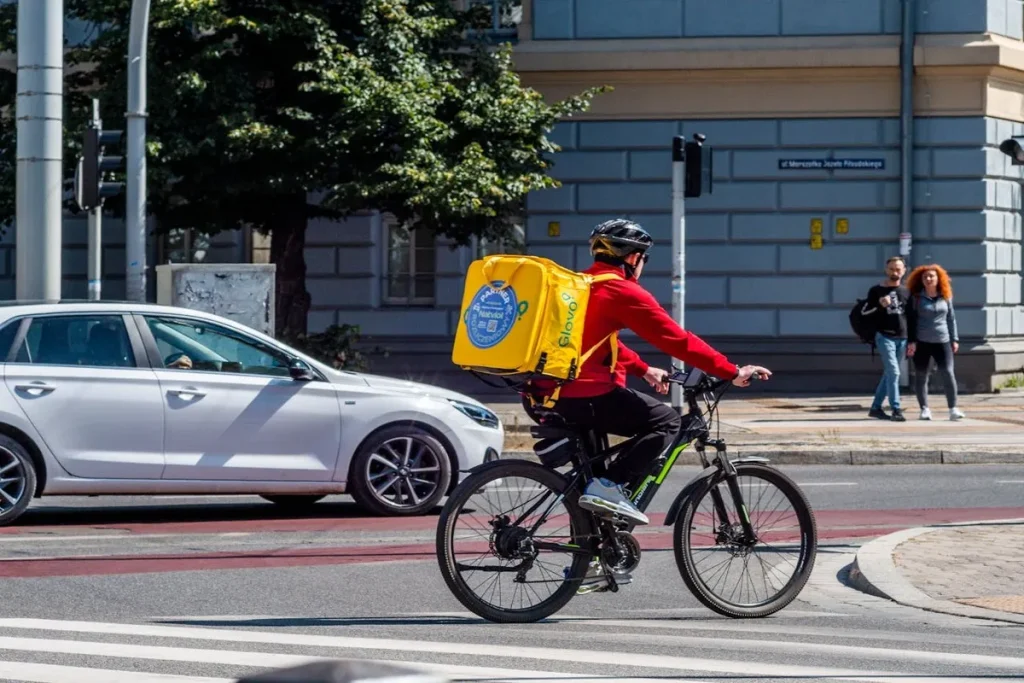 Um entregador de aplicativo, vestindo uma jaqueta vermelha, capacete e uma mochila amarela grande nas costas, está pedalando uma bicicleta na cidade. A imagem representa o trabalho de profissionais autônomos, como entregadores de aplicativo.