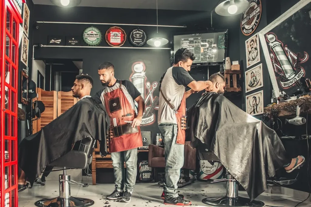 Interior de uma barbearia com dois barbeiros atendendo clientes. Eles usam aventais de couro e estão focados no corte de cabelo. A imagem representa o trabalho de profissionais autônomos e o ambiente de uma barbearia.