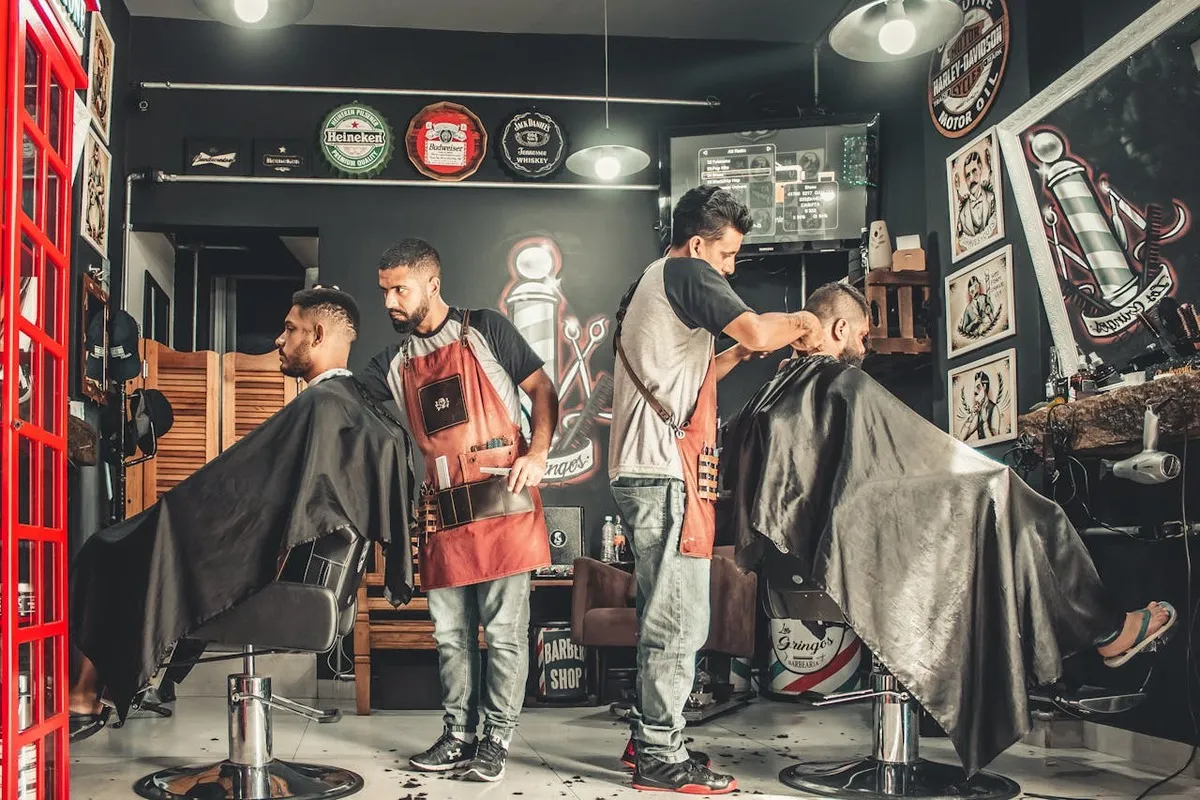 Interior de uma barbearia com dois barbeiros atendendo clientes. Eles usam aventais de couro e estão focados no corte de cabelo. A imagem representa o trabalho de profissionais autônomos e o ambiente de uma barbearia.