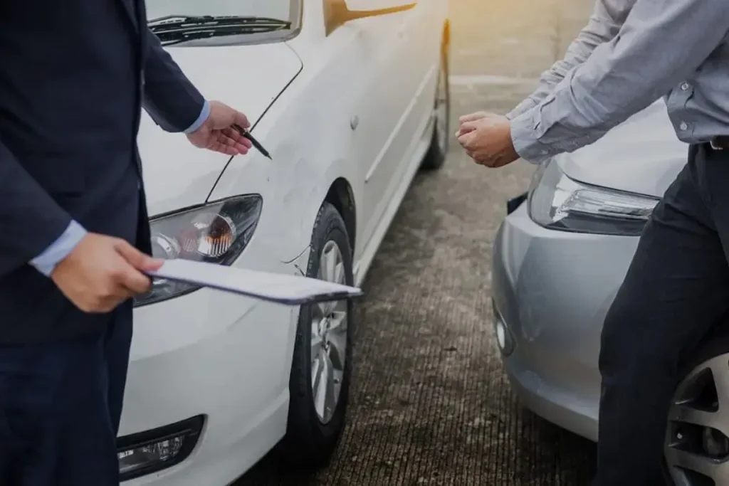 Um homem de terno e outro de camisa social examinam os danos de dois carros brancos após uma colisão. A imagem simboliza a busca e apreensão de veículos.