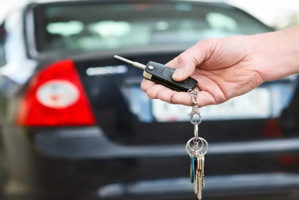 Mão entregando ou pegando chave de carro em frente à traseira de um veículo escuro, simbolizando a busca e apreensão por falta de pagamento.