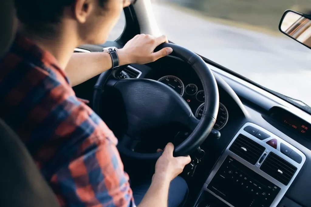 Homem dirigindo um carro, mostrando as mãos no volante e o painel do veículo.
