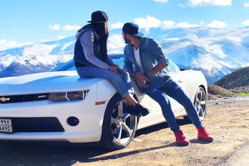 Um casal sentado em um carro Chevrolet Camaro branco, com montanhas nevadas no fundo.