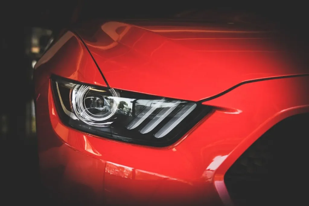 Close-up do farol e da parte frontal de um carro esportivo vermelho, provavelmente um Ford Mustang.