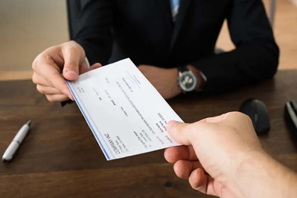 Uma pessoa de terno entrega um cheque a outra pessoa, que o recebe com a mão estendida. As mãos estão em destaque no centro da imagem, com uma caneta e um mouse de computador no fundo desfocado. A imagem simboliza pagamento, transação financeira ou a entrega de crédito.