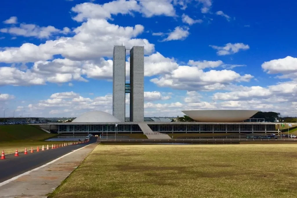 Vista externa do Congresso Nacional em Brasília, com suas duas torres verticais e as cúpulas do Senado e da Câmara dos Deputados, sob um céu azul com nuvens brancas. A imagem representa o cenário de deliberações e aprovações de leis que podem influenciar a criação e a gestão de um consórcio público.
