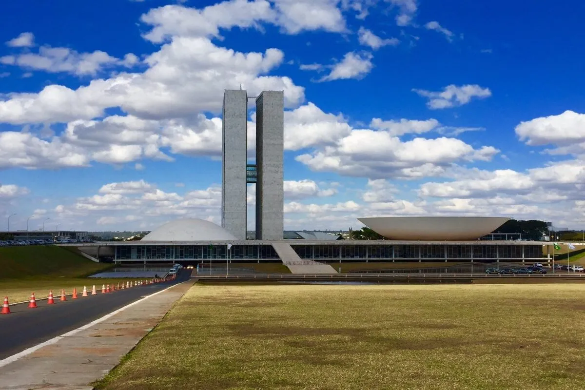 Vista externa do Congresso Nacional em Brasília, com suas duas torres verticais e as cúpulas do Senado e da Câmara dos Deputados, sob um céu azul com nuvens brancas. A imagem representa o cenário de deliberações e aprovações de leis que podem influenciar a criação e a gestão de um consórcio público.