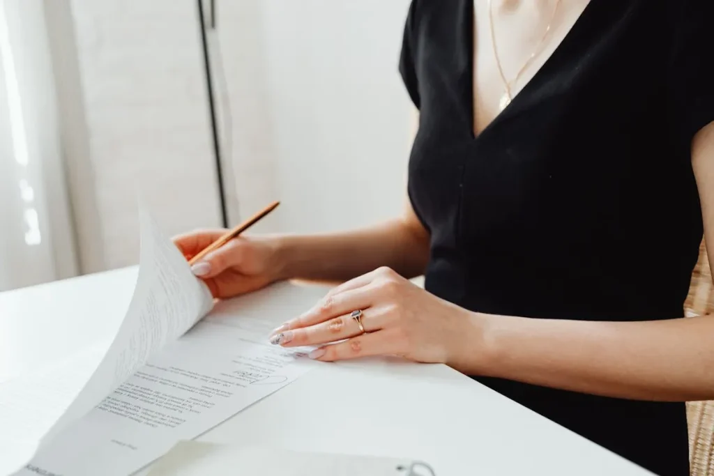 Uma mulher, usando um anel e um colar, analisa cuidadosamente um documento ou contrato sobre uma mesa. A imagem representa o estudo das cláusulas e responsabilidades, como as de um devedor solidário, antes de assinar.
