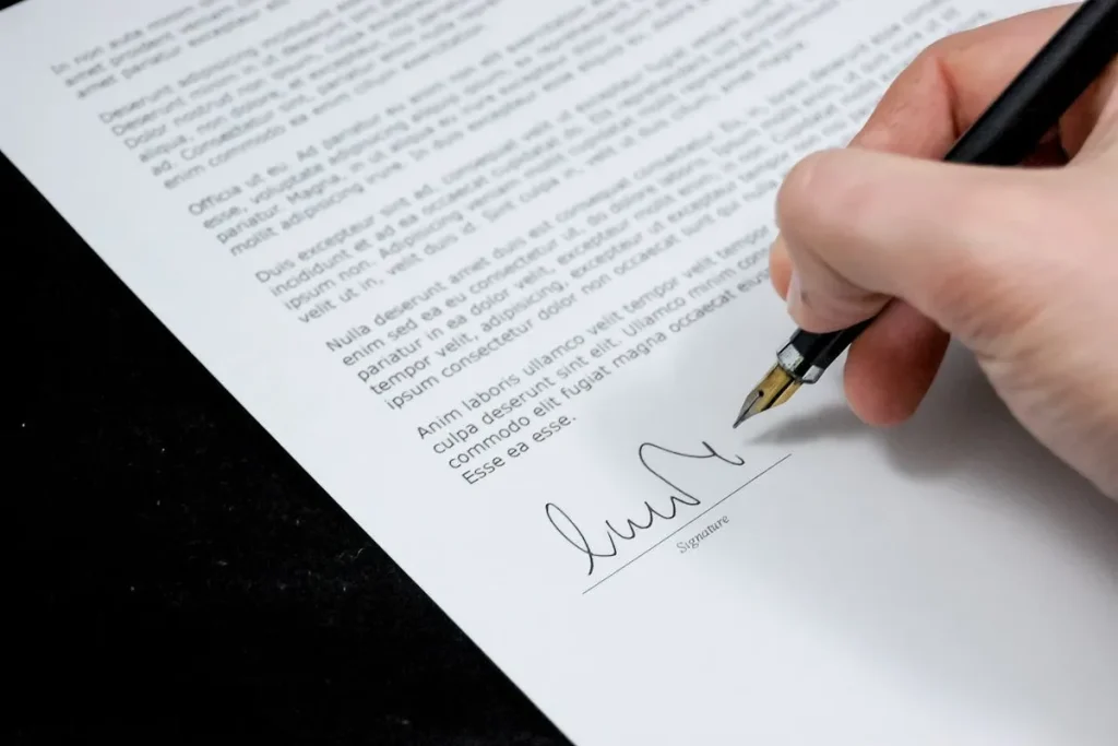 Uma pessoa assina um documento com uma caneta-tinteiro, com o foco na assinatura e no texto legal do contrato. A imagem simboliza a formalização de um compromisso financeiro, como o de um devedor solidário.