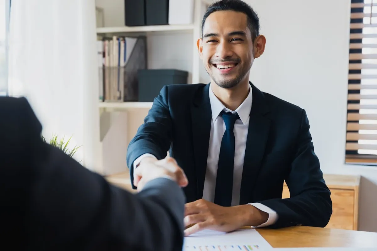 Dois homens de negócios, um deles sorrindo, apertam as mãos sobre uma mesa de escritório com documentos e gráficos. A imagem representa a conclusão de um acordo, possivelmente envolvendo um devedor solidário.