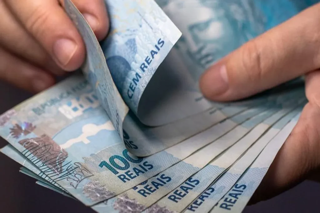 Uma pessoa contando notas de 100 reais, com uma mão segurando as notas e a outra ajeitando as mesmas. A imagem representa o tema de dinheiro, finanças e o tema da lei do consórcio devolução do dinheiro 2024.