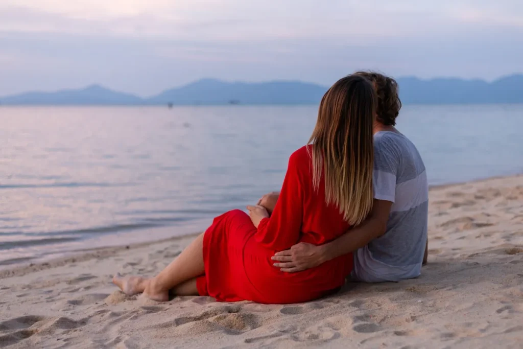 Um casal sentado de costas na areia de uma praia, abraçados e observando o mar e as montanhas ao entardecer, ilustrando a palavra-chave 'lugares para passar lua de mel'.
