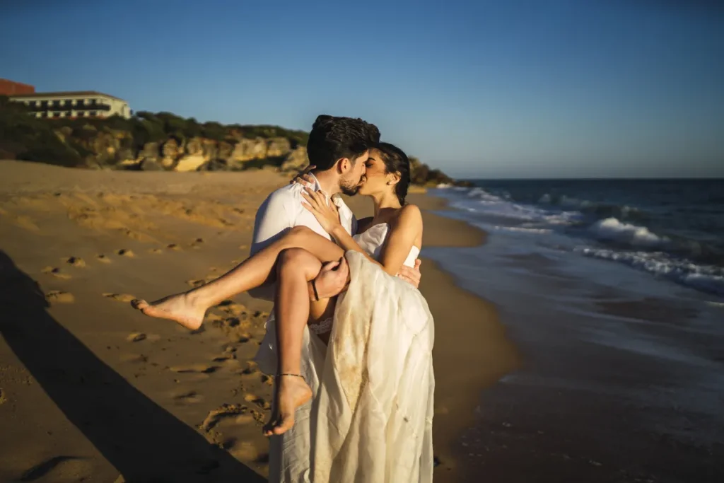 Um casal de noivos em um abraço romântico, com o homem segurando a mulher no colo na areia de uma praia ao entardecer, ilustrando a palavra-chave 'lugares para passar lua de mel'.