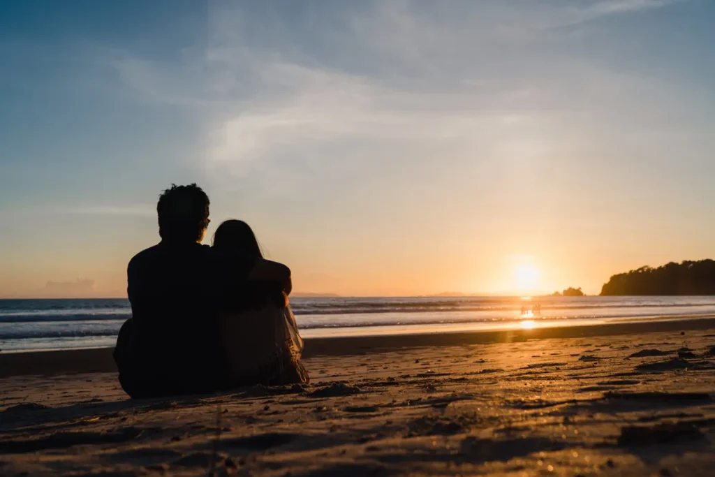 Um casal sentado na areia da praia, abraçado, observando o pôr do sol sobre o mar, ilustrando a palavra-chave 'lugares para passar lua de mel' e viagens românticas.