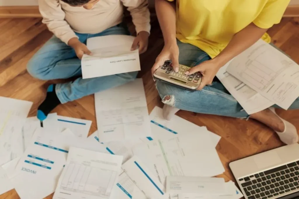 Vista superior de duas pessoas, uma adulta e uma criança, sentadas no chão rodeadas por contas e uma calculadora, representando a organização financeira para evitar o nome sujo.