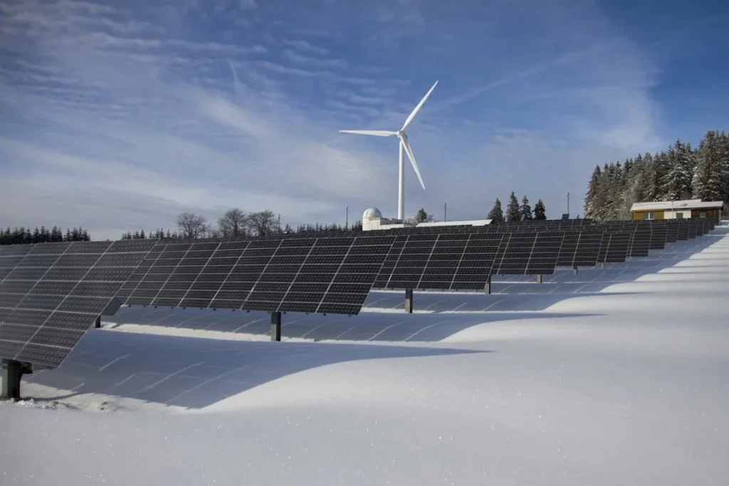 Painéis solares em uma área coberta de neve, com um moinho de vento e árvores ao fundo sob um céu azul.
