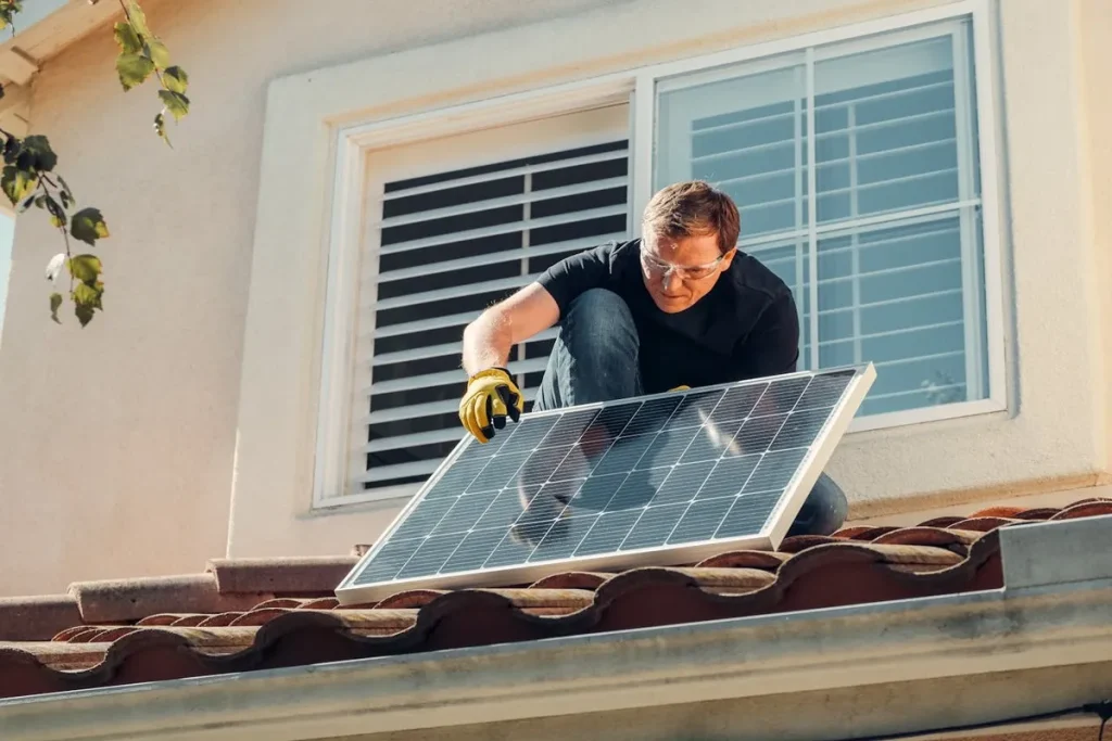 Homem instalando um dos painéis solares em telhado de telhas, usando óculos de proteção e luvas, com uma casa ao fundo.