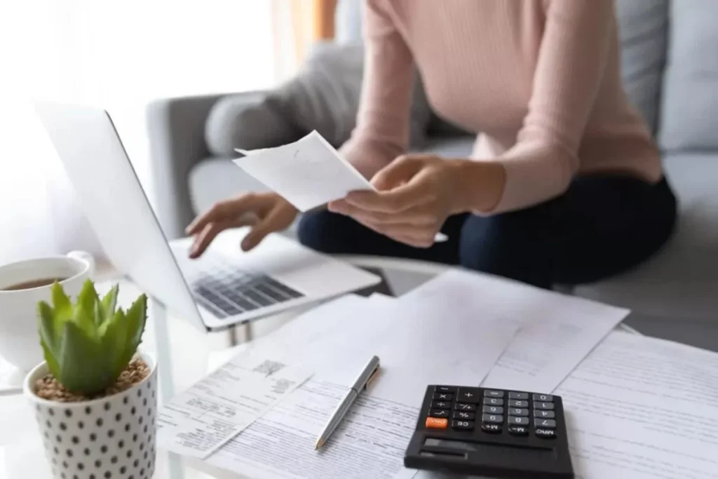 Mulher sentada em casa, revisando documentos e usando uma calculadora e um laptop. A imagem representa a gestão financeira pessoal e o cálculo de despesas de forma 'pro rata', como o rateio de contas e juros.