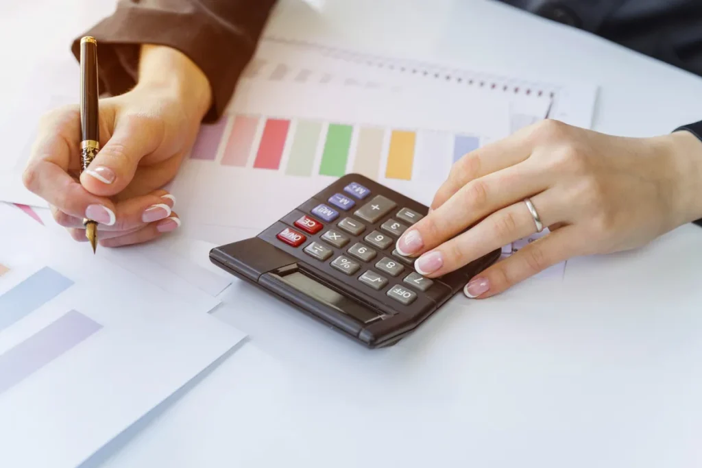 Mãos de uma pessoa utilizando uma calculadora e segurando uma caneta sobre papéis e gráficos, em uma mesa de escritório. A imagem representa o ato de fazer cálculos financeiros e a análise de dados para determinar valores de forma 'pro rata'.