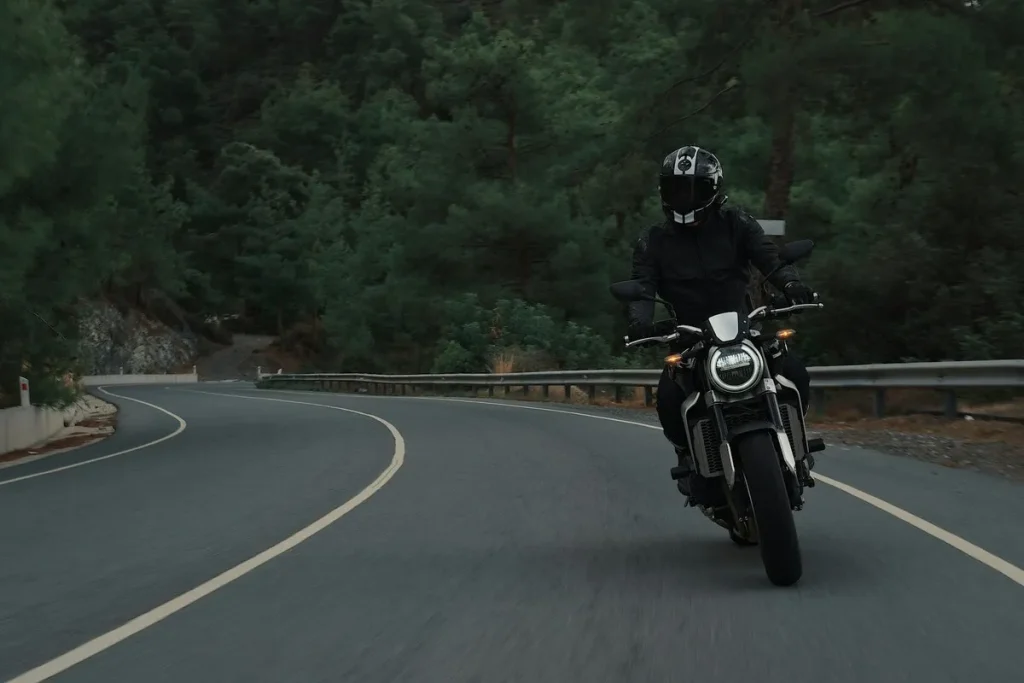 Motociclista usando jaqueta de couro e capacete preto, pilotando uma moto esportiva branca em uma estrada sinuosa cercada por floresta.
