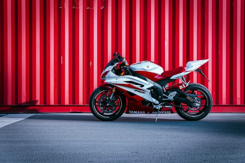 Uma motocicleta esportiva Yamaha R6, branca e vermelha, estacionada em frente a uma parede vermelha com listras verticais.