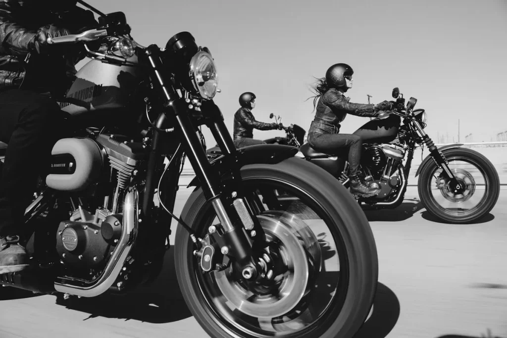 Imagem em preto e branco de três motociclistas, sendo duas mulheres, pilotando motocicletas Harley-Davidson em uma estrada.